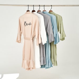 May include: Five silk robes hanging on a rail, in a row. The robes are in shades of blush, white, grey, blue, and green. The blush robe has the word "Bride" printed on the back. Each robe has a belt and ruffled sleeves.