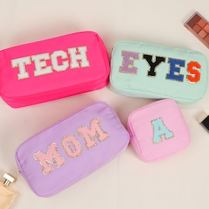 May include: Four colorful cosmetic bags with text. A hot pink bag says "TECH", a mint green bag says "EYES", a lavender bag says "MOM", and a pink bag says "A". Makeup and perfume bottles are in the background.