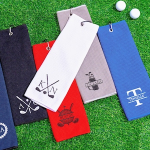Puede incluir: Toallas de golf en varios colores, incluyendo azul, marino, rojo, blanco y gris, se muestran sobre una superficie verde. Cada toalla presenta un diseño personalizado diferente, como iniciales, nombres o gráficos temáticos de golf. También se ven pelotas de golf.
