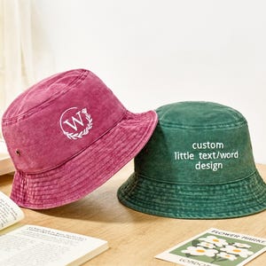 May include: A magenta bucket hat featuring a white letter W inside a circular wreath design and a dark green bucket hat with the white text custom little text/word design embroidered on the front, both made of a washed-out fabric material.