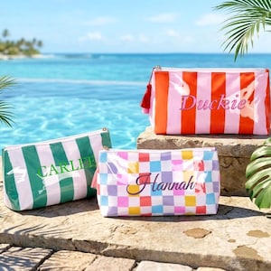 Peut inclure: Trois trousses rectangulaires à fermeture éclair avec des prénoms brodés sur le devant. Une trousse présente des rayures verticales vertes et blanches avec le nom Carlee, une autre des rayures orange et roses avec Duckie, et une troisième un motif à damier multicolore avec Hannah.