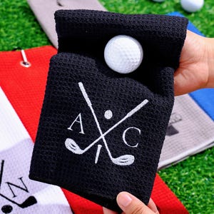 May include: A black golf towel with a white golf ball inside. The towel has a white embroidered design of crossed golf clubs and the letters "A" and "C". Other golf towels are visible in the background.
