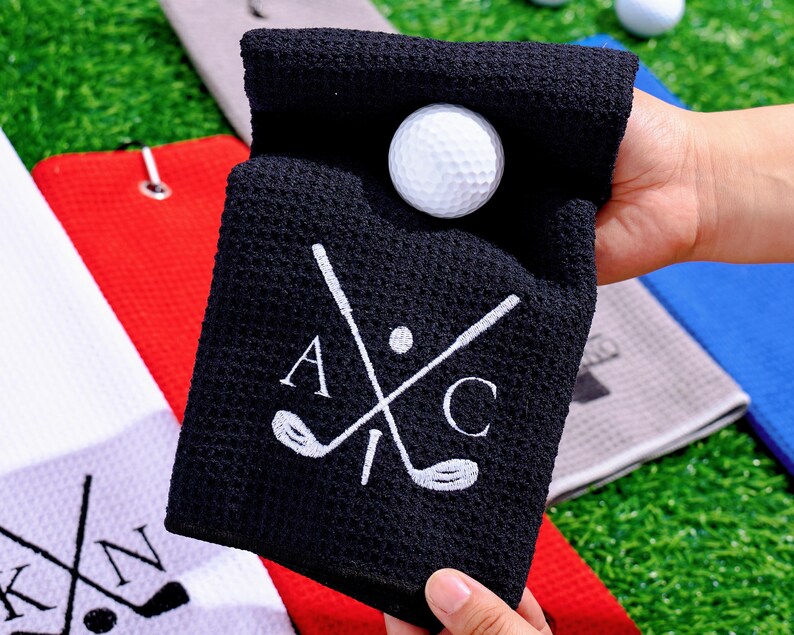 May include: A black golf towel with a white golf ball inside. The towel has a white embroidered design of crossed golf clubs and the letters "A" and "C". Other golf towels are visible in the background.