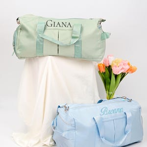 Puede incluir: Dos bolsas de viaje personalizadas, una verde claro con el nombre "Giana" y otra azul claro con el nombre "Breana". Las bolsas est&aacute;n sobre una superficie blanca con un ramo de tulipanes rosas y amarillos en un jarr&oacute;n.