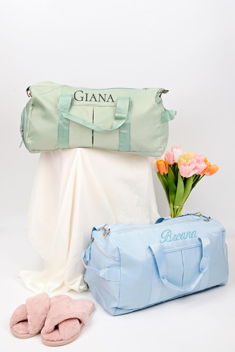 Puede incluir: Dos bolsas de viaje personalizadas, una verde claro con el nombre "Giana" y otra azul claro con el nombre "Breana". Las bolsas est&aacute;n sobre una superficie blanca con un ramo de tulipanes rosas y amarillos en un jarr&oacute;n.