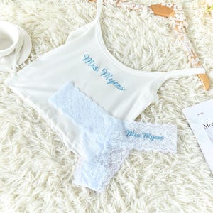 May include: White lingerie set featuring a camisole and a thong. The camisole and thong are embroidered with "Mrs. Myers" in light blue script. The thong is made of light blue lace.