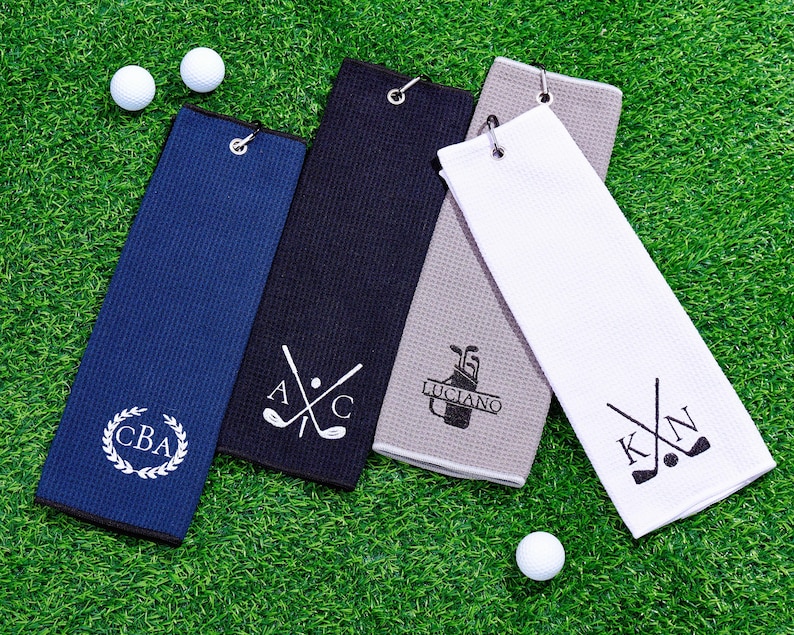 May include: Four golf towels in navy blue, black, gray, and white, each with a different design and personalized initials. Two golf balls are visible on the green grass. The towels have a loop for hanging.