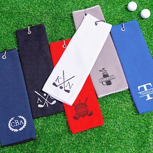 May include: Golf towels in various colors, including blue, navy, red, white, and gray, are arranged on a green surface. Each towel features a different personalized design, such as initials, names, or golf-related graphics. Golf balls are also visible.