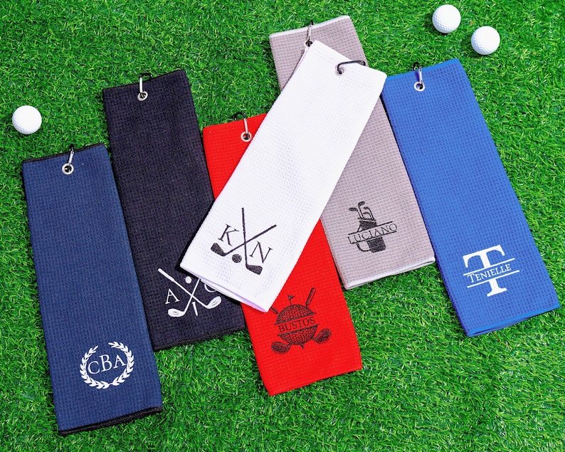 May include: Golf towels in various colors, including blue, navy, red, white, and gray, are arranged on a green surface. Each towel features a different personalized design, such as initials, names, or golf-related graphics. Golf balls are also visible.