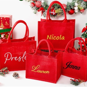May include: A collection of red tote bags with handles, each personalized with a name in a contrasting color. The bags are displayed with Christmas decorations, including a small tree and gift boxes, suggesting a holiday theme.