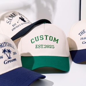 May include: A collection of baseball caps with off-white crowns and contrasting brims in navy blue or green. The caps feature embroidered text and graphics, including the words CUSTOM EST.2025, MITCHELL, and SAME HOLE, along with palm tree and golfer icons.