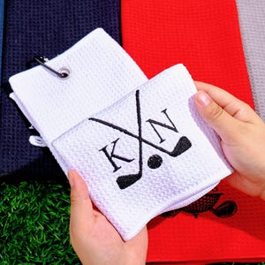 May include: White golf towel with a waffle weave texture. The towel features crossed golf clubs, a golf ball, and the initials "KN" embroidered in black. A silver clip is attached to the towel. Other colored towels are visible in the background.