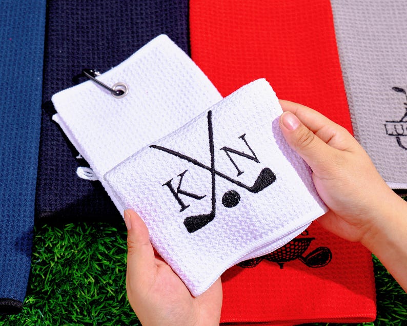May include: White golf towel with a waffle weave texture. The towel features crossed golf clubs, a golf ball, and the initials "KN" embroidered in black. A silver clip is attached to the towel. Other colored towels are visible in the background.