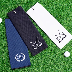 May include: Three golf towels in navy blue, black, and white, with golf ball accents. The towels feature personalized initials and golf club designs. The towels are made of a textured material and have a metal grommet for hanging.