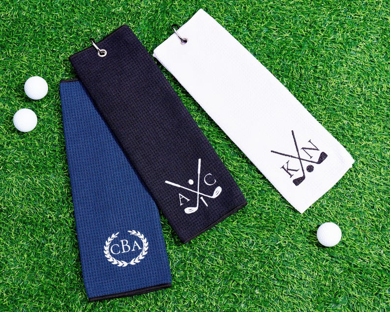 May include: Three golf towels in navy blue, black, and white, with golf ball accents. The towels feature personalized initials and golf club designs. The towels are made of a textured material and have a metal grommet for hanging.