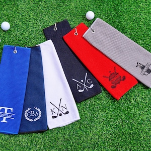 May include: Golf towels in blue, navy, white, red, and gray, with golf ball accents. Each towel features a different embroidered design, including initials, crossed golf clubs, and the name "Bustos" and "Luciano". The towels are on a green surface.