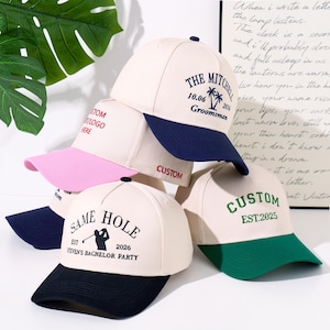 May include: A collection of five baseball caps with cream-coloured crowns and contrasting brims in navy blue, pink, and green. The caps feature embroidered text and graphics, including "THE MITCHELLS," "SAME HOLE," "CUSTOM," and "STEVEN'S BACHELOR PARTY."