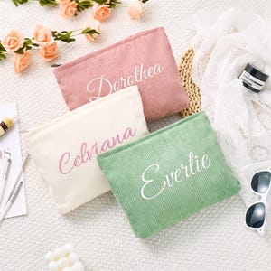 Puede incluir: Tres estuches rectangulares de pana en rosa empolvado, crema y verde salvia. Cada estuche cuenta con cierre de cremallera y un nombre bordado en letra cursiva: Dorothea en el rosa, Celviana en el crema y Everlie en el verde.