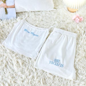 Puede incluir: Conjunto de pijama blanco con camiseta de tirantes y pantalones cortos. La camiseta de tirantes tiene el texto "Mrs. Myers" en escritura azul. Los pantalones cortos tienen el texto "EST 10/22/25" en escritura azul. El conjunto se muestra sobre una alfombra blanca y esponjosa.