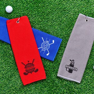 May include: Golf towels in red, blue, and gray, each with a personalized design and a metal grommet for hanging. The red towel features the name "Bustos", the blue towel has "Taylor", and the gray towel displays "Luciano". Two golf balls are also visible.