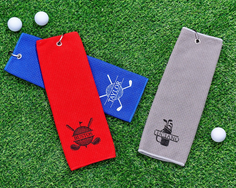 May include: Golf towels in red, blue, and gray, each with a personalized design and a metal grommet for hanging. The red towel features the name "Bustos", the blue towel has "Taylor", and the gray towel displays "Luciano". Two golf balls are also visible.