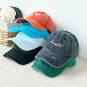 May include: A stack of baseball caps in various colors, including orange, blue, and black. The orange cap has "CY" embroidered on the front, while a gray cap displays the word "Beautiful". The caps are made of a soft fabric.