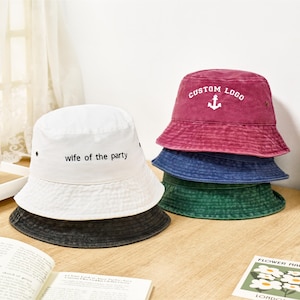 May include: A white bucket hat with the text "wife of the party" printed in black, stacked atop a dark grey bucket hat. Beside it, a stack of three distressed bucket hats in maroon, blue, and green, with the top maroon hat featuring an anchor logo.