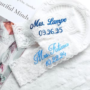 May include: Two white lace garters with blue embroidered text. One reads "Mrs. Lampe 09.26.25" and the other "Mrs. Fatima 10.22.24". The garters are displayed on a white, decorative plate.