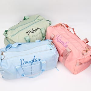 Puede incluir: Tres bolsas de viaje personalizadas en diferentes colores. Una bolsa azul claro con el nombre "Douglas" bordado en azul, una bolsa verde claro con el nombre "Mehan" bordado en negro y una bolsa rosa con el nombre "Rambe" bordado en morado.