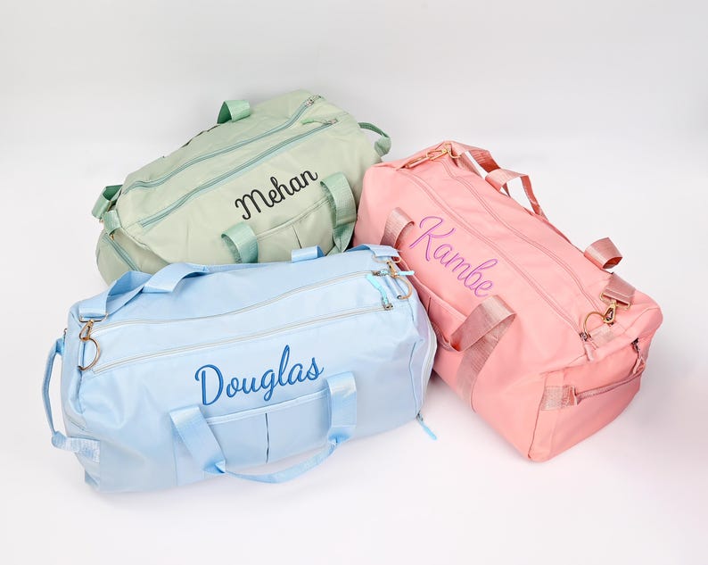 Puede incluir: Tres bolsas de viaje personalizadas en diferentes colores. Una bolsa azul claro con el nombre "Douglas" bordado en azul, una bolsa verde claro con el nombre "Mehan" bordado en negro y una bolsa rosa con el nombre "Rambe" bordado en morado.