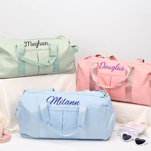 Puede incluir: Tres bolsas de viaje personalizadas en diferentes colores. La primera bolsa es verde claro con el nombre "Meghan" bordado. La segunda bolsa es rosa con el nombre "Douglas" bordado. La tercera bolsa es azul claro con el nombre "Milan" bordado.