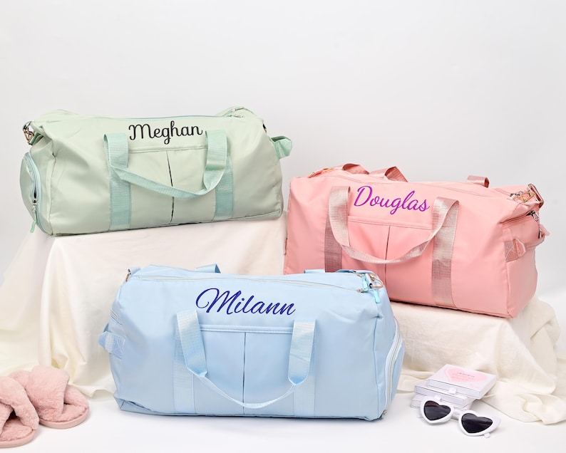 Puede incluir: Tres bolsas de viaje personalizadas en diferentes colores. La primera bolsa es verde claro con el nombre "Meghan" bordado. La segunda bolsa es rosa con el nombre "Douglas" bordado. La tercera bolsa es azul claro con el nombre "Milan" bordado.