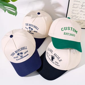 May include: Four baseball caps with off-white crowns and coloured brims. Two caps feature navy brims with blue text reading THE MITCHELL 10.06 2026 Groom and Groomsman. One cap has a green brim with green text CUSTOM EST.2025. One cap has a black brim with black text.