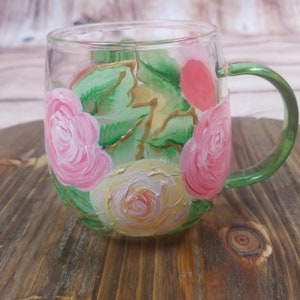 May include: A clear glass mug with a green handle, hand-painted with pink, yellow, and white roses and green leaves.