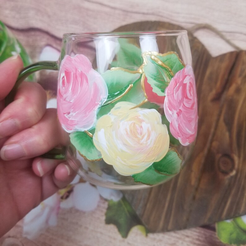 May include: A clear glass mug with a hand-painted design of pink and yellow roses with green leaves. The roses are painted in a realistic style with delicate details.