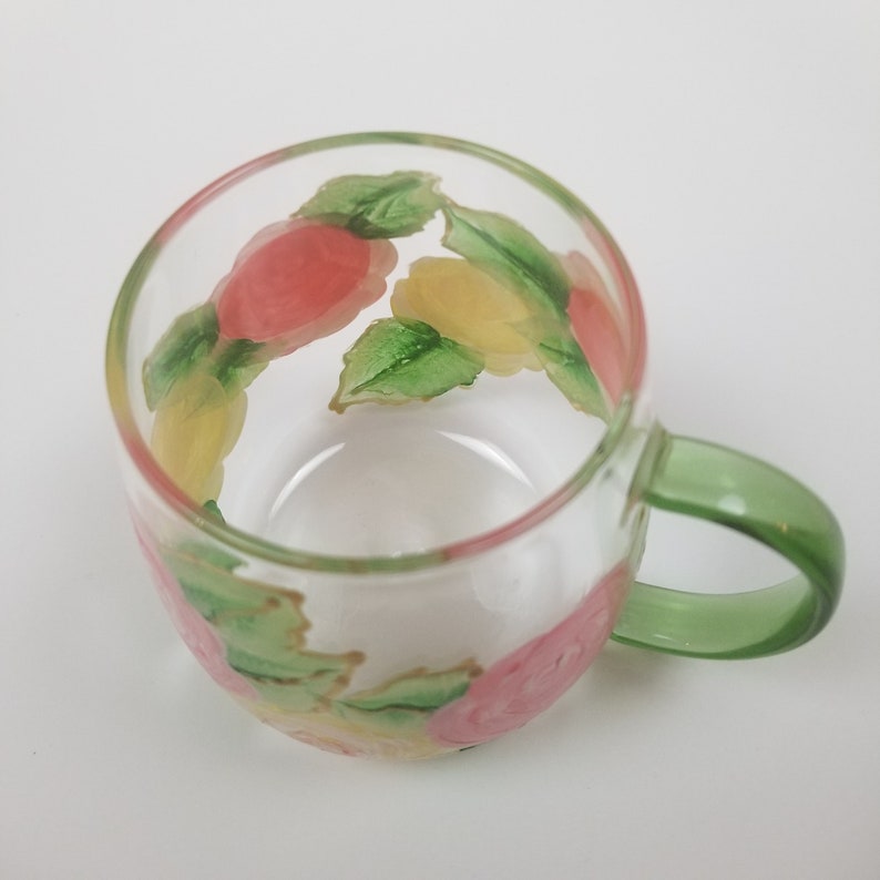 May include: A clear glass mug with a green handle. The mug is decorated with hand-painted pink, yellow, and green roses.