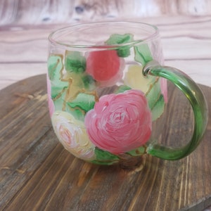May include: A clear glass mug with a green handle, hand-painted with pink, yellow, and white roses and green leaves.
