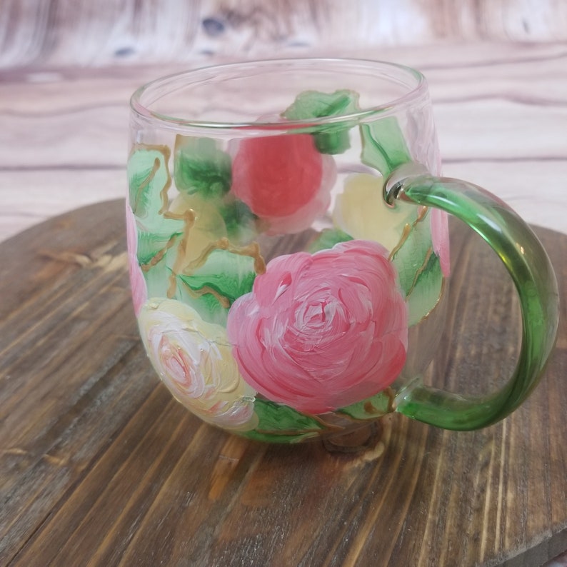 May include: A clear glass mug with a green handle, hand-painted with pink, yellow, and white roses and green leaves.