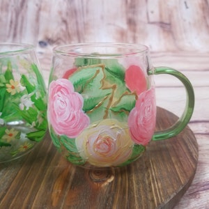 May include: A hand-painted glass mug with a green handle. The mug features a floral design with pink and yellow roses, green leaves, and gold accents.