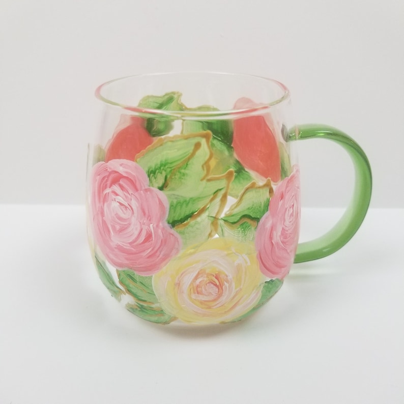 May include: A glass mug with a green handle, hand-painted with pink and yellow roses and green leaves.