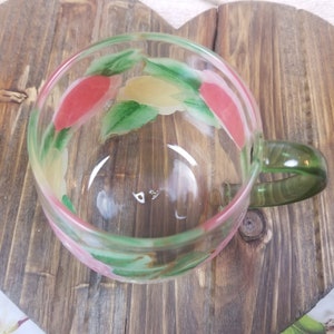 May include: A clear glass mug with a green handle. The mug has a floral design painted on the outside with pink, yellow, and green colors.