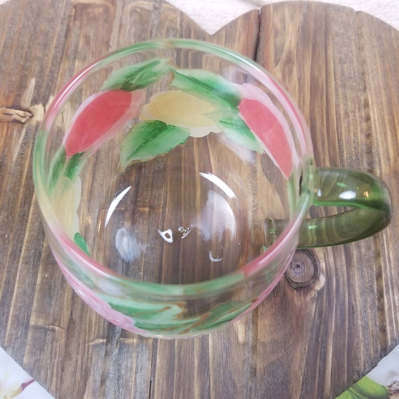 May include: A clear glass mug with a green handle. The mug has a floral design painted on the outside with pink, yellow, and green colors.