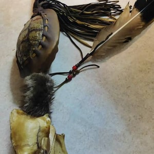 May include: A Native American ceremonial rattle with a skull, feathers, and leather. The rattle is decorated with red beads and has a brown leather handle.