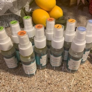 May include: A collection of ten small glass spray bottles with white caps and colorful labels. The bottles are filled with a clear liquid and are arranged in a row on a table. The labels on the bottles have different colors and text, including "Refreshing Room Spray" and "Wild Orange".