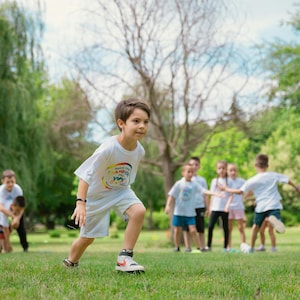 Può includere: Un gruppo di bambini che gioca all'aperto su un campo erboso. Un bambino indossa una maglietta bianca con un design colorato e corre verso la fotocamera. Gli altri bambini sono in piedi o camminano sullo sfondo.