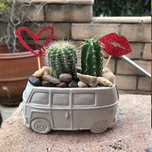 May include: A gray concrete Volkswagen bus planter with two small cacti and red glitter hearts and lips.