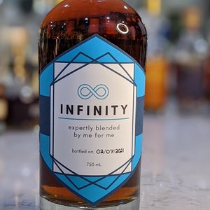 May include: A clear glass bottle with a wooden cork, filled with amber liquid. The label reads "INFINITY" with a blue and white geometric design. Text on the label includes "expertly blended by me for me" and "bottled on: 02/07/2021". The bottle contains 750 ml.