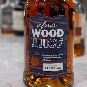 May include: A bottle of amber liquid with a label reading "infinite WOOD JUICE." The label features a wood grain design and the text "100% Natural Pulp Free Goodness." The bottle is dated 07/27/20.