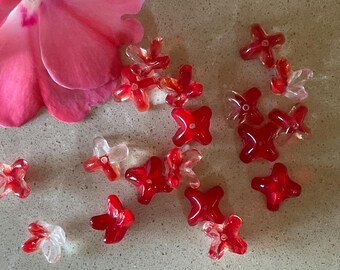 May include: A collection of red and clear, star-shaped beads scattered on a light-colored surface, with a pink flower petal in the upper left corner. The beads vary in transparency and shade of red.