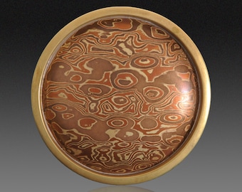 May include: A round, decorative metal plate with a gold-colored rim. The center features a swirling pattern of brown, copper, and cream tones. The design resembles a topographical map or the surface of a planet. The plate is likely used for decorative purposes.